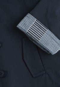 Manteau marine présentant un motif pied de poule texturé sur le poignet, avec une coupe ajustée et un détail de poche frontale.