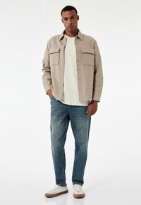 Chaqueta de camisa beige con botones y dos bolsillos en el pecho, llevada sobre una camiseta blanca, combinada con jeans azules y zapatillas blancas con suelas en tono marrón.