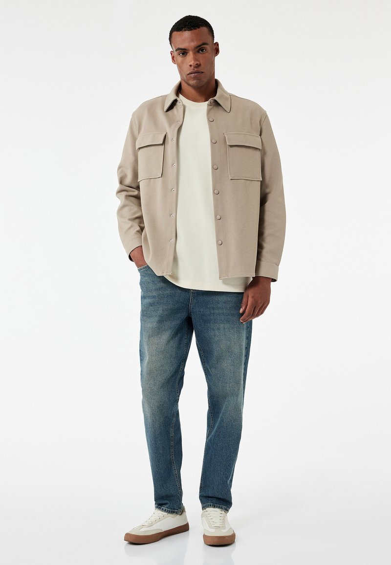 Chaqueta de camisa beige con botones y dos bolsillos en el pecho, llevada sobre una camiseta blanca, combinada con jeans azules y zapatillas blancas con suelas en tono marrón.