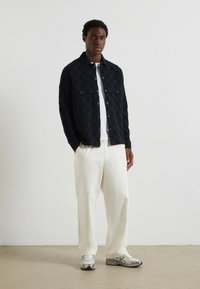 Giacca a scacchi blu navy con bottoni, dotata di due tasche frontali, abbinata a pantaloni larghi beige e sneakers grigie, su uno sfondo neutro.
