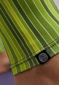 Zielone legginsy w paski wykonane z elastycznej tkaniny, z pionowymi liniami w różnych odcieniach zieleni oraz małą czarną metką z logo w kształcie spirali.