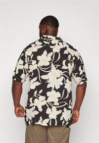 Camisa de manga corta de tela negra con un gran patrón floral en color crema. Presenta un cuello y un ajuste relajado con aberturas laterales.