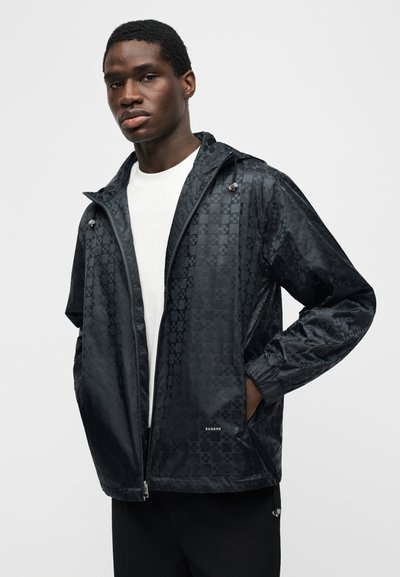 Νέος άνδρας που φοράει μαύρο καρό μπουφάν με φερμουάρ και κουκούλα πάνω από λευκό πουκάμισο, όρθιος με το ένα χέρι στην τσέπη, σε απλό φόντο.