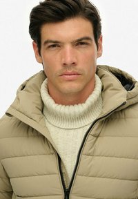 Giovane uomo con capelli scuri che indossa una giacca imbottita beige sopra un maglione a collo alto bianco, rivolto in avanti con un'espressione neutra.