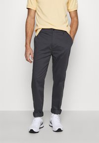 Man wearing beige short-sleeve shirt, dark gray rolled-up pants, and white sneakers standing against a plain light background.