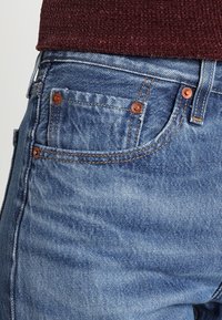 Pantalones vaqueros de mezclilla azul claro con remaches de color cobre, diseño clásico de cinco bolsillos y textura ligeramente descolorida.