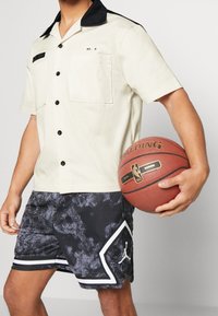 Camicia a maniche corte color crema con colletto nero, abbinata a pantaloni corti neri a fantasia con dettagli bianchi e logo, mentre tiene un pallone da basket.