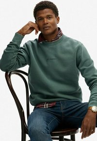 Sudadera verde con logotipo bordado, llevada sobre una camisa de cuadros rojos, combinada con jeans azules. Modelo sentado en una silla de madera.