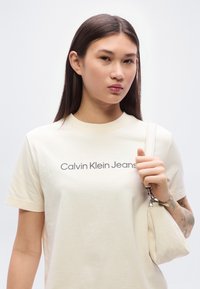 Μπεζ βαμβακερό μπλουζάκι με εκτυπωμένο λογότυπο "Calvin Klein Jeans" σε σκούρο γκρι, συνδυασμένο με μια μικρή τσάντα χειρός σε αντίστοιχη απόχρωση.