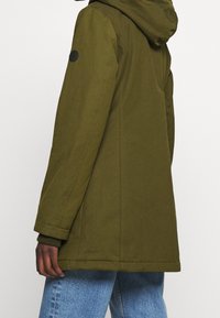 Person iført en olivengrøn hættejakke med ribbede manchetter og lyseblå jeans, vist fra ryggen og siden.