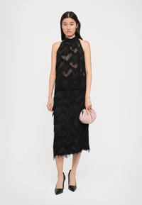 Svart kjole med lagdelte frynser og halterneck. Har gjennomsiktig bodice og en tettsittende silhuett. Et par svarte stiletthæler og en rosa veske.