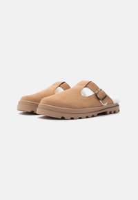 Barna velúr slip-on cipők, puha fehér béléssel, oldalsó csattal és tartós gumitalppal, amelynek barázdázott mintázata van.