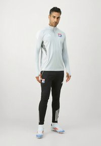 Hellgraues Trainingsoberteil mit Reißverschluss, kombiniert mit schwarzen, akzentuierten langen Hosen und weißen Streifen. Mit einem "OL"-Logo auf der Brust und Fußballschuhen.