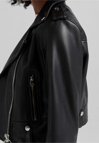 Musta keinonahkainen moottoripyörätakki, jossa on hopeanväriset vetoketjut, painonapit ja vinosti sijoitettu taskudetalji. Takissa on istuva, lyhyt siluetti.