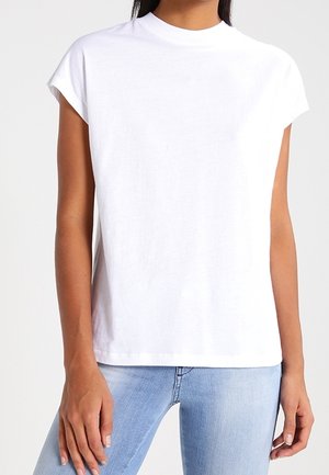 Femme portant un t-shirt blanc uni à manches courtes et col rond, ainsi qu'un jean bleu clair, debout avec les bras détendus.