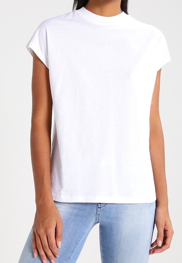 Femme portant un t-shirt blanc uni à manches courtes et col rond, ainsi qu'un jean bleu clair, debout avec les bras détendus.