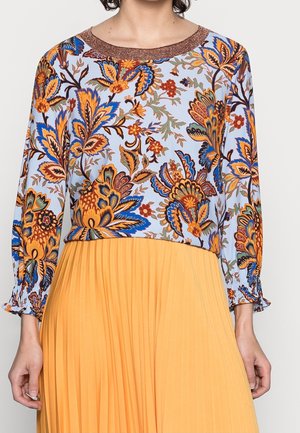 Vrouw draagt een blouse met lange mouwen met blauwe, oranje en bruine bloemenpatronen, gecombineerd met een gele, geplooide rok met hoge taille.