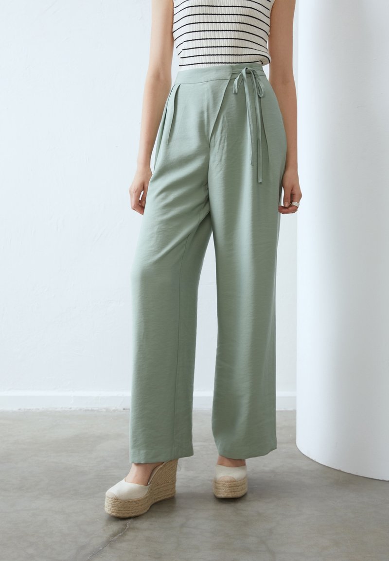 Personne portant un pantalon large taille haute vert clair avec une ceinture nouée, un haut sans manches rayé et des chaussures compensées beiges, debout à l'intérieur.