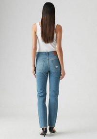 Jean bleu ajusté en denim avec une taille mi-haute, jambes droites, décoloration subtile et une pièce en cuir marron au dos. Porté avec des talons noirs.