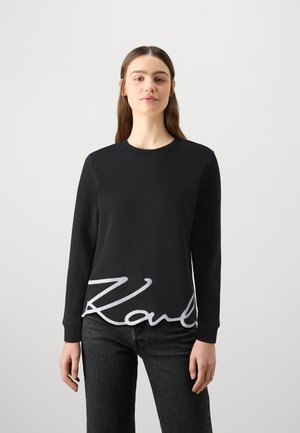 Schwarzer Sweatshirt mit langen Ärmeln und rundem Halsausschnitt. Verfügt über einen großen weißen Signaturdruck am Saum. Weiches Material und einfaches Design.
