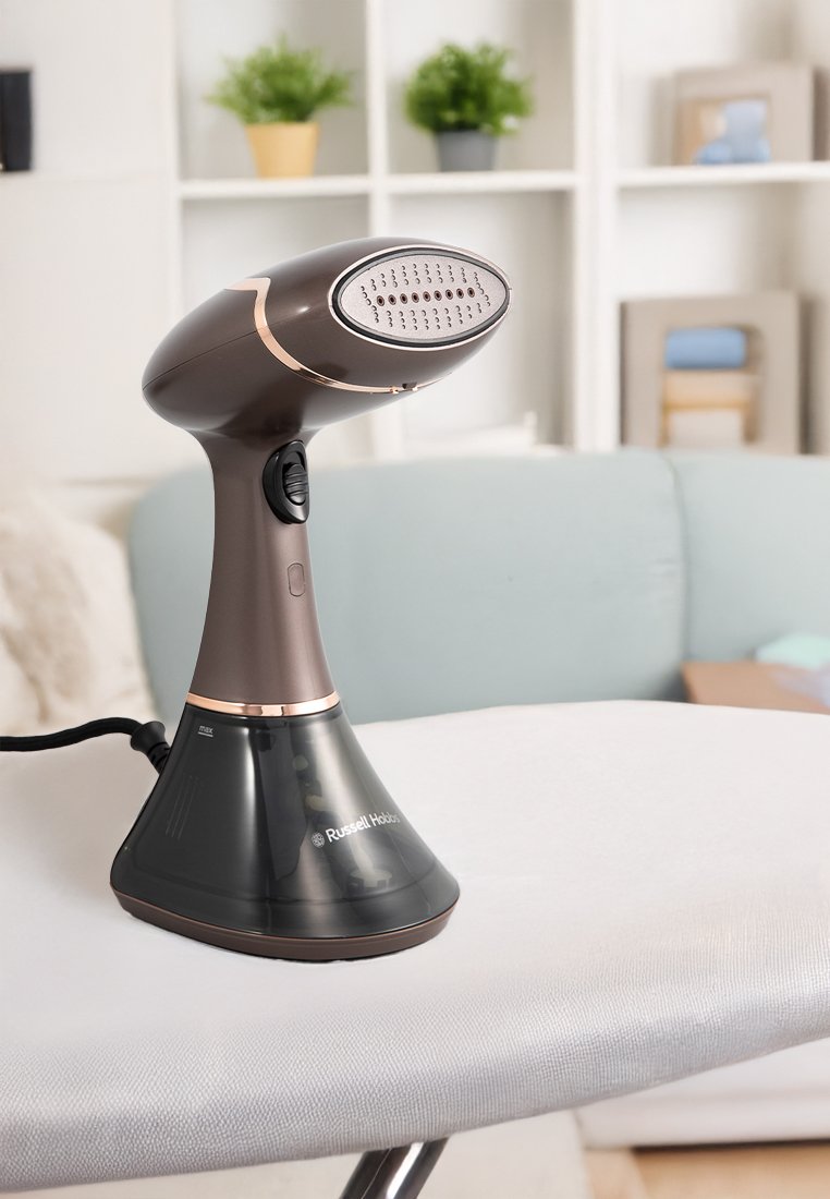 Vaporizador de prendas portátil en marrón y negro sobre una tabla de planchar, con estanterías y plantas de salón desenfocadas al fondo.