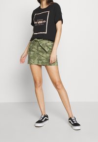 Svart kortärmad t-shirt med vit och roséguld Vans-logotyp, i kombination med en grön kamouflagekjol med en rosett vid midjan och svarta sneakers.