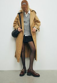 Vaaleanruskea trench-takki, harmaa neuleliivi, valkoinen paita, mustat hameet, läpikuultavat sukkahousut, ruskeat loafer-kengät kultaisilla yksityiskohdilla, musta kudottu laukku, baseball-lippis.