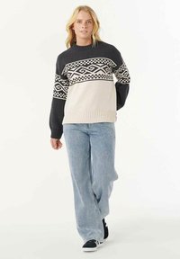 Una donna bionda che cammina, indossando un maglione lavorato a maglia a motivi bianchi e neri, jeans a vita alta di un blu chiaro e scarpe da ginnastica nere.