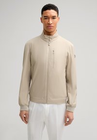 Beige lichtgewicht jack met een hoge kraag, een ritssluiting aan de voorkant en elastische manchetten. Beschikt over een ritsborstzak en een zachte textuur.