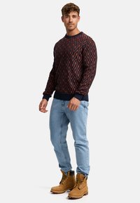 Pulover cu model în roșu închis și bleumarin, cu design în diamante, asociat cu blugi de culoare bleu deschis și cizme cu șireturi de culoare bej. Mâneci și tiv ribate.