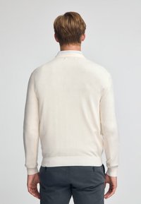 Kremowy sweter o miękkiej fakturze, z prążkowanymi mankietami i dołem oraz klasycznym okrągłym dekoltem. Nosi się go na szarych spodniach.