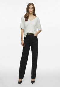Maglia bianca a V in cotone abbinata a pantaloni neri a vita alta e tacchi neri. L'outfit presenta un punto vita segnato da una cintura e un fit rilassato.