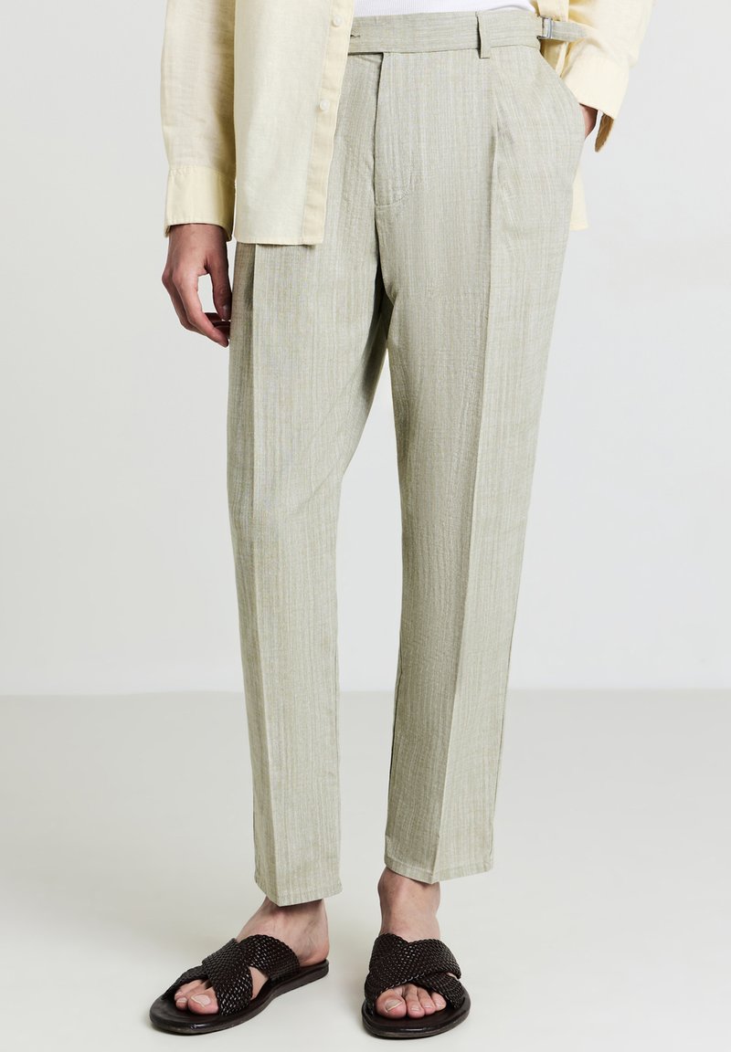 Pantaloni beige a vita alta con sottili righe, abbinati a sandali intrecciati marrone scuro e una camicia crema con bottoni.