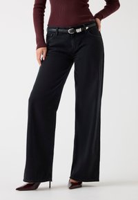 Sorte viddejeans lavet af denim, med et sort bælte med sølvfarvet hardware og et højtaljet design. Glat tekstur, ingen synlige mønstre.