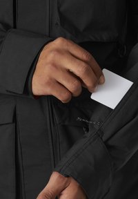 Chaqueta negra con una textura suave, que cuenta con un bolsillo lateral donde se está insertando una tarjeta blanca. Costuras visibles y puños ajustables.