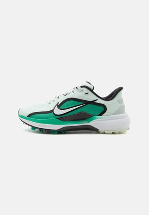 Weißer und grüner Sportschuh mit schwarzen Akzenten, Nike-Logo und profilierter Sohle mit Spikes, seitlich fotografiert vor weißem Hintergrund.