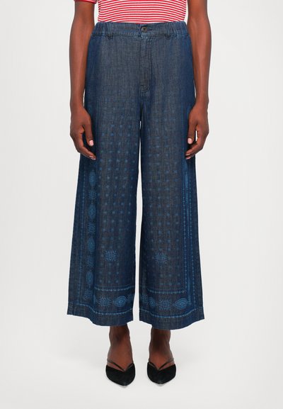 Pantaloni in denim a gamba larga di colore blu scuro con un design a motivo. Presentano una chiusura con bottone e una consistenza liscia, abbinati a scarpe nere.