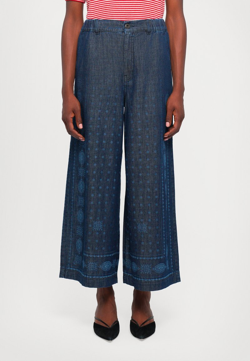Pantaloni in denim a gamba larga di colore blu scuro con un design a motivo. Presentano una chiusura con bottone e una consistenza liscia, abbinati a scarpe nere.