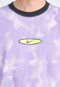 Purpursvars T-krekls ar apaļu Nike logotipu zaļā krāsā. Audumam ir teksturēts, punktots raksts ar kontrastējošu melnu apkakli.