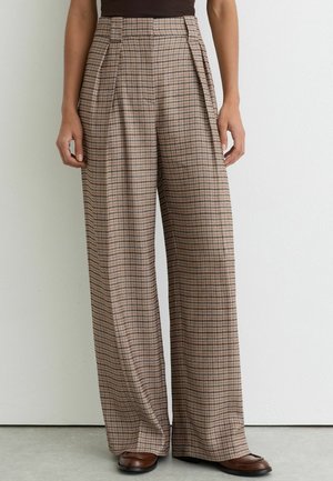 Žena nosiaca kárované nohavice s vysokým pásom a širokými nohavicami v béžových a hnedých tónoch, doplnené hnedými topánkami, stojaca pred obyčajnou bielou stenou.