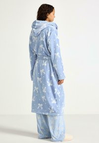 Niebieski szlafrok z polaru z kapturem, ozdobiony białym wzorem w kokardki i kropki. Noszony na dopasowanych niebieskich spodniach do spania z podobnym wzorem. Tkanina o fakturze.