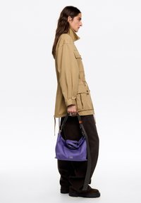 Violetti käsilaukku, jossa on sileä pinta, varustettu suljetulla ylhäällä, kontrastivärillä varustetulla hihnalla ja logodetaililla. Yhdistetty vaaleanruskeaan takkiin ja tummiin housuihin.