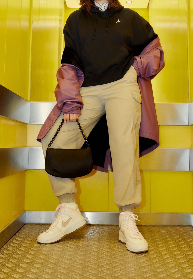 Personne portant un pantalon beige, des baskets blanches, un sweat à capuche noir, une veste violette, tenant un sac à main noir, debout dans un ascenseur aux murs jaunes.