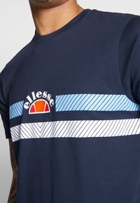 Námořnické tričko s krátkým rukávem vyrobené z bavlny, s logem a světle modrými a bílými diagonálními pruhy přes hrudník.