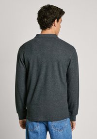 Polo a maniche lunghe di colore grigio scuro realizzato in tessuto a coste, con colletto, fessure laterali e vestibilità rilassata, abbinato a jeans azzurri.
