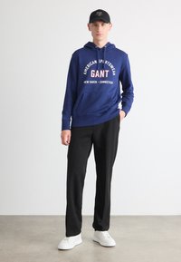 Sudadera azul con el logo "GANT" y bolsillo frontal, combinada con pantalones negros y zapatillas blancas, complementada por una gorra negra. Textura suave.