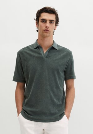 Młody mężczyzna o ciemnych kręconych włosach, ubrany w zieloną koszulę z krótkim rękawem i kołnierzykiem oraz białe spodnie, ręce w kieszeniach, neutralne tło.