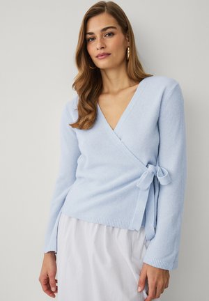 Kvinde med langt, bølget hår iført guldringe i ørerne og en lyseblå omslagsstriktrøje bundet i siden, kombineret med et hvidt skørt.