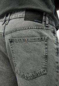 Gros plan sur la poche arrière d'un jean en denim gris et une étiquette de marque noire sur la ceinture, porté avec une chemise sombre.