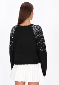 Fekete pulóver bolyhos textúrával, amelyet flitterek díszítenek a vállakon, fehér pliszírozott szoknyával párosítva.